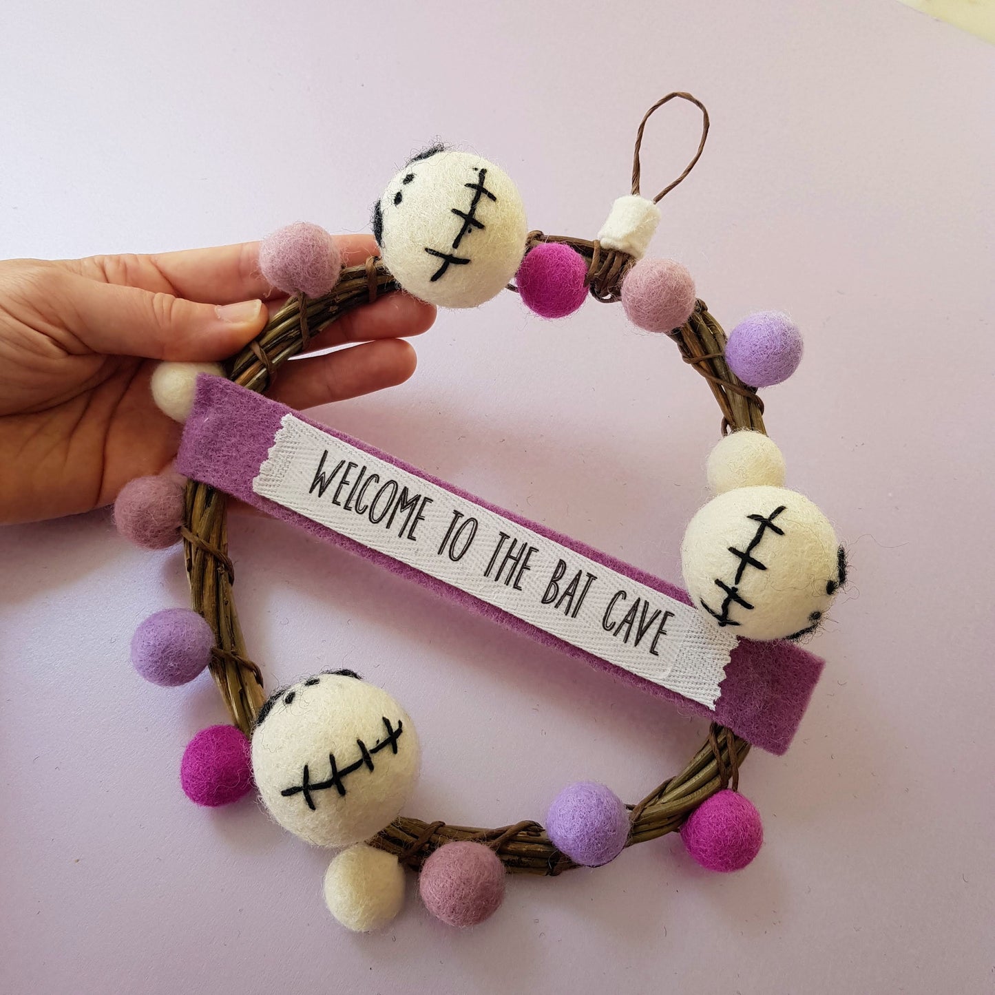 Decorative wreath with pom-poms and a sign saying 'Welcome to the Bat Cave' on a light pink background.