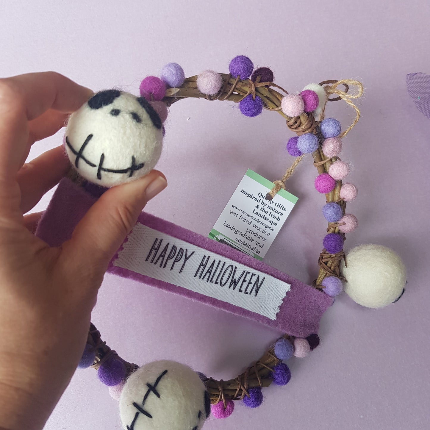 Decorative wreath with Halloween-themed pom-poms and a 'Happy Halloween' ribbon on a light purple background.