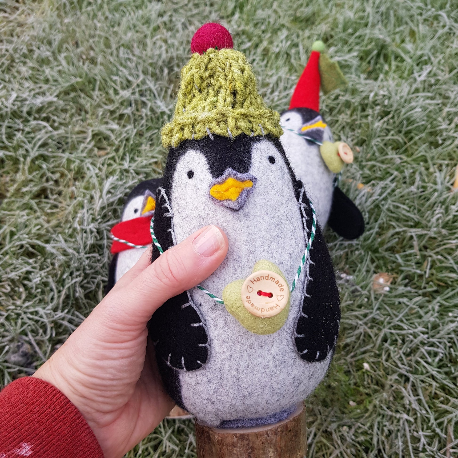 Hand holding a felt penguin ornament with a green hat against a grassy background