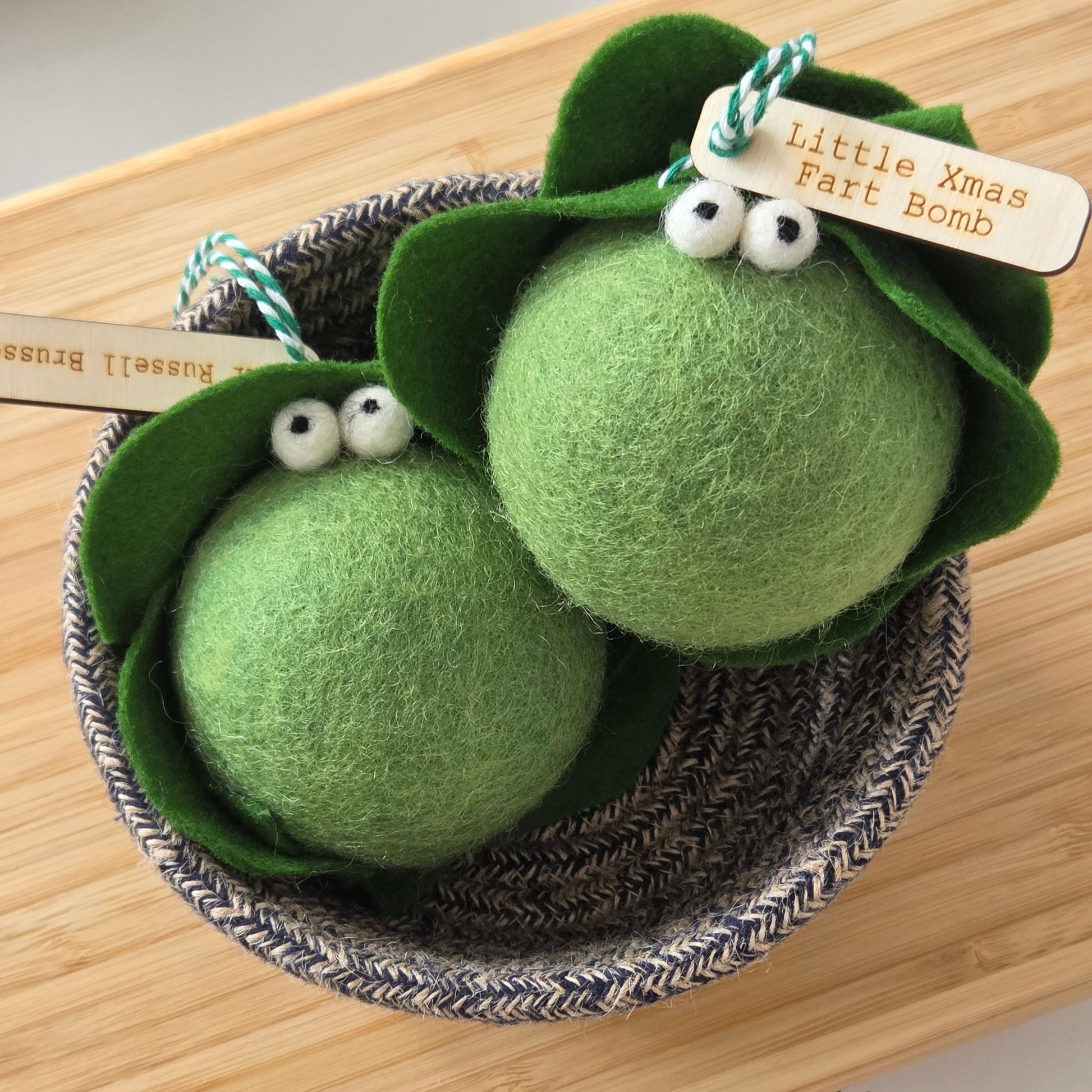 Two green felted Christmas ornaments shaped like peas in a basket on a wooden surface with pine cones.