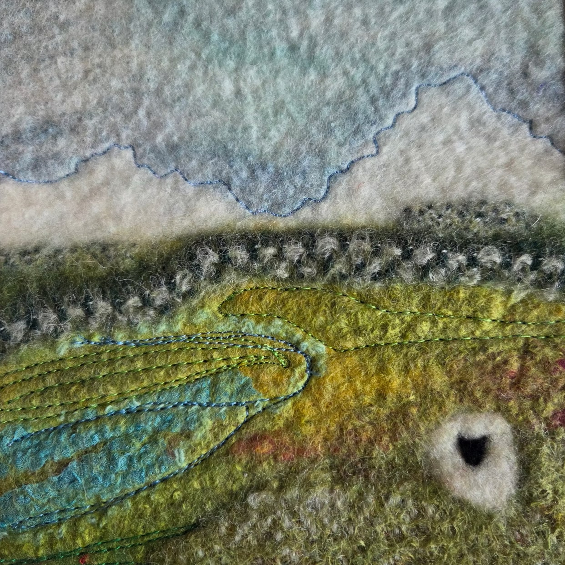 Textile art piece with a landscape design featuring green fields and a blue sky.