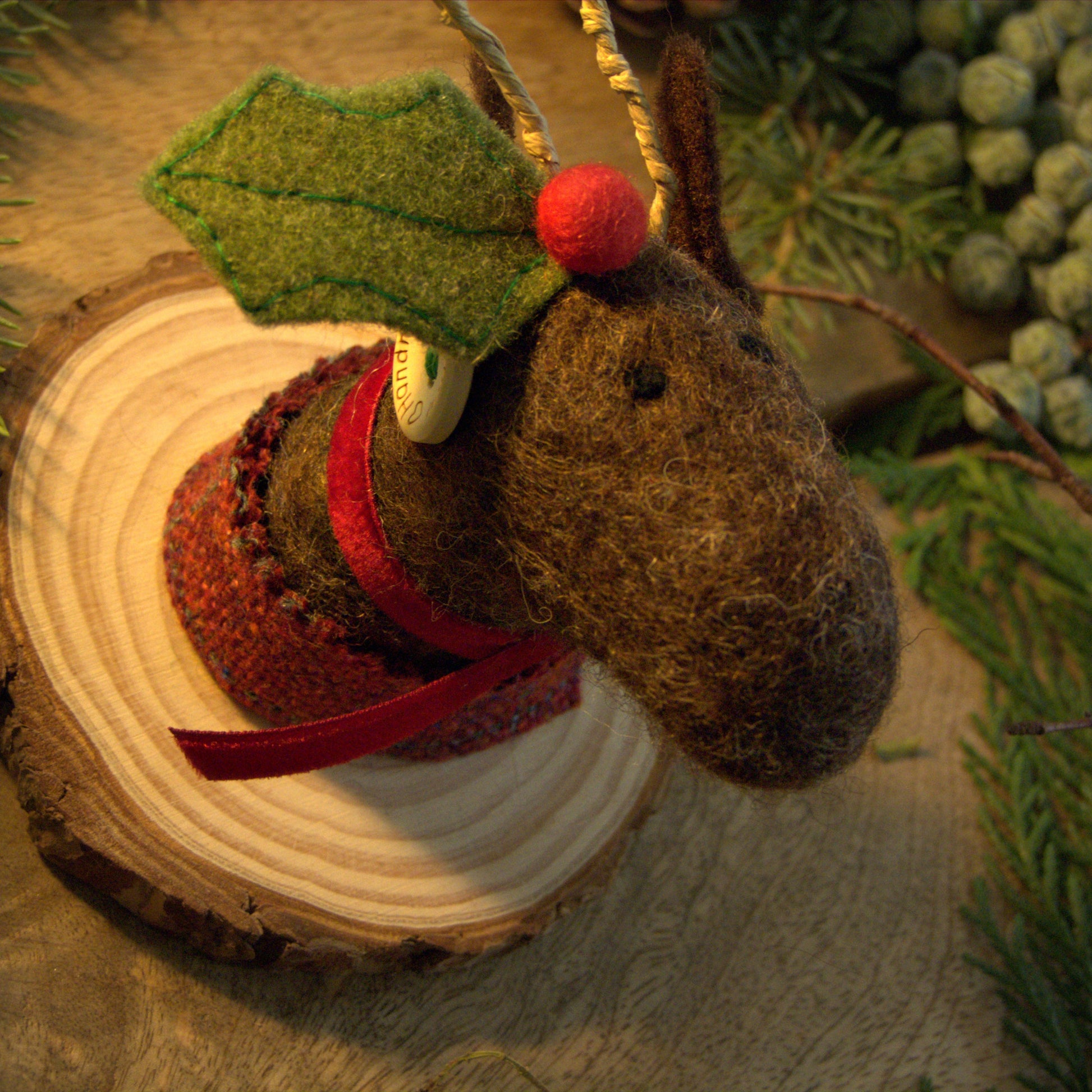 Felted reindeer ornament with holly leaf and red nose on a wooden log with greenery.