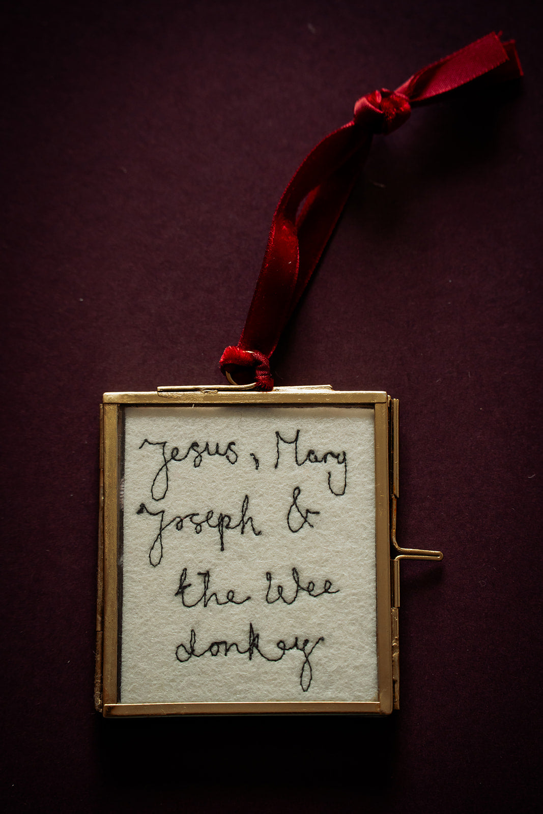 A square framed ornament with a red ribbon loop, featuring embroidered black cursive text on a cream fabric background, set against a dark burgundy backdrop.