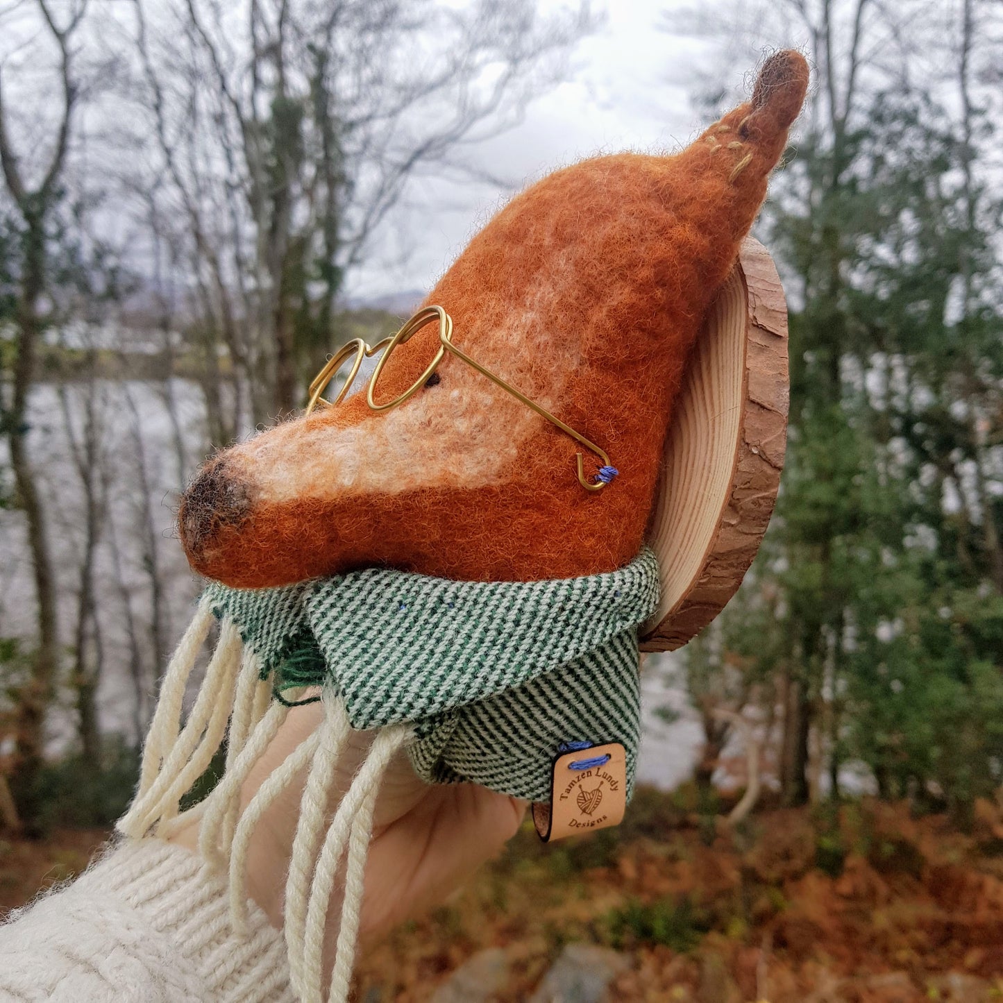 Felted animal sculpture of a fox with a scarf, held outdoors with trees and water in the background.