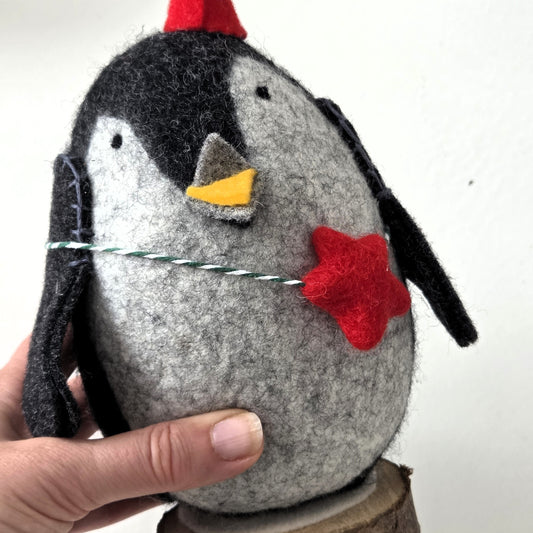 Handmade penguin ornament with a red hat and star, held by a hand against a white background.