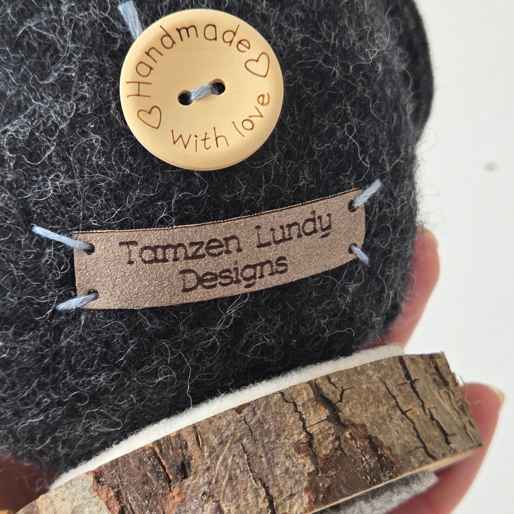 Close-up of a textured fabric item with a gold button and 'Tanzen Lundy Designs' label, held by a hand.