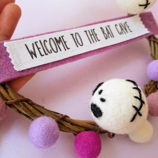 Decorative wreath with a plush toy and 'Welcome to the Bat Cave' sign on a pink background