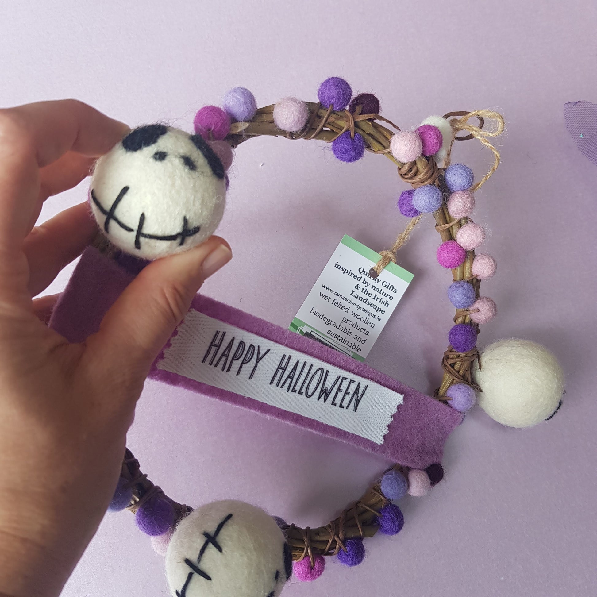 Decorative wreath with Halloween-themed pom-poms and a 'Happy Halloween' ribbon on a light purple background.