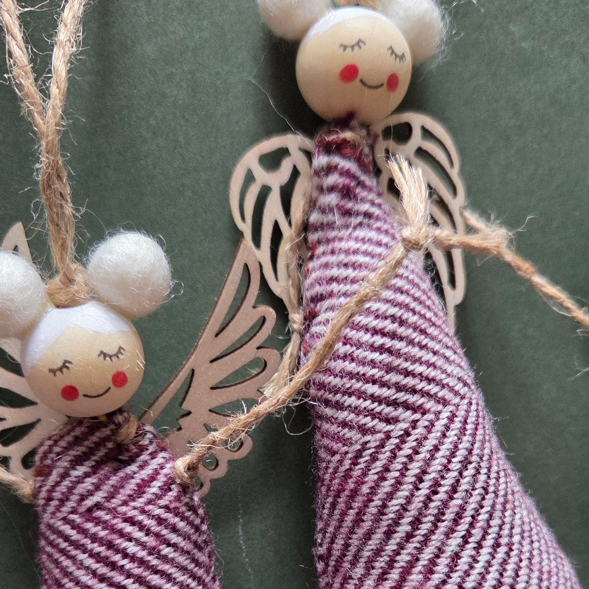 Two handmade fabric angel ornaments with striped purple and white cloaks, wooden faces, and white pom-pom hair, hanging against a dark green background.