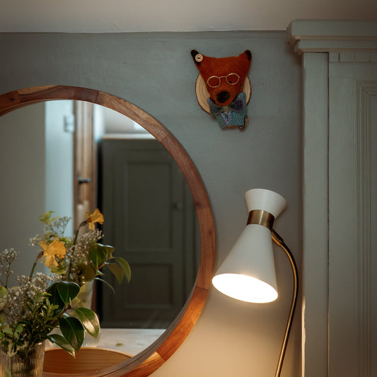 Round wooden mirror on a table with a lamp and decorative fox head on the wall.