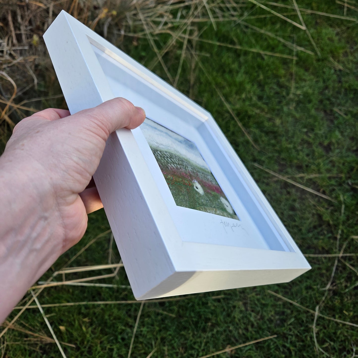 Hand holding a white framed picture against a grassy background