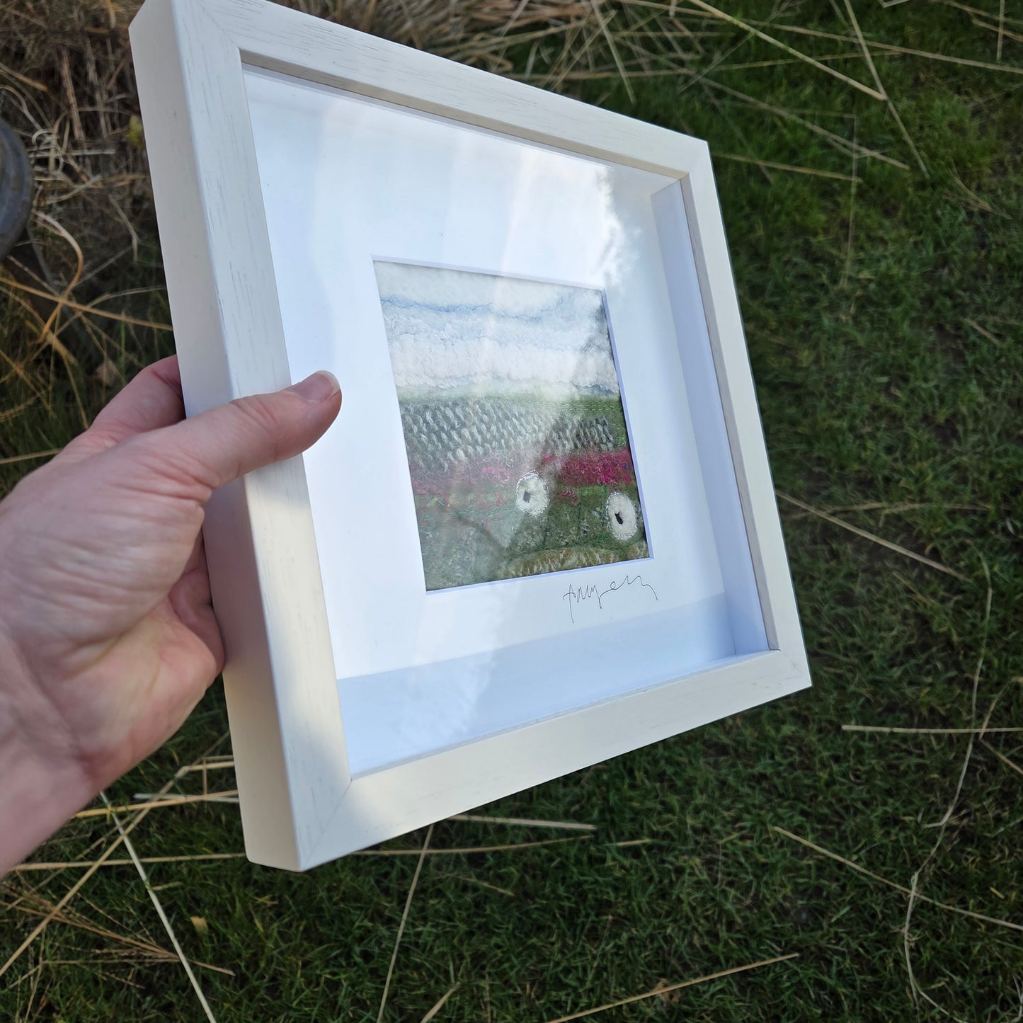 Hand holding a white framed picture against a grassy background
