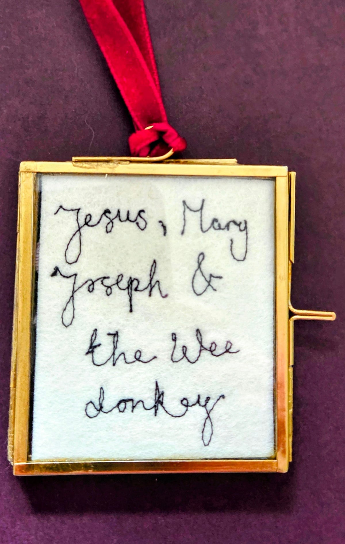 A small square gold-framed pendant or ornament with white fabric insert and handwritten black text reading 'Jesus, Mary, Joseph & the wee donkey', hanging from a red ribbon on a dark purple background.
