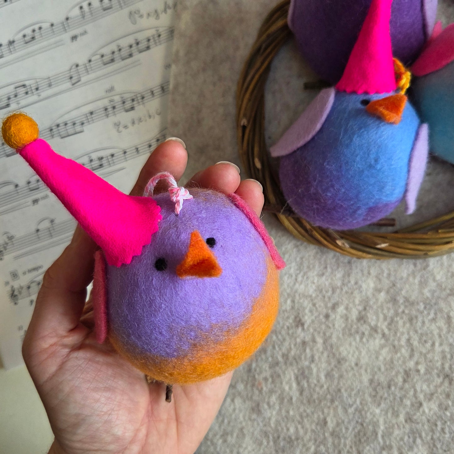 Colorful felt bird toy held in a hand with more toys in the background on a textured surface.