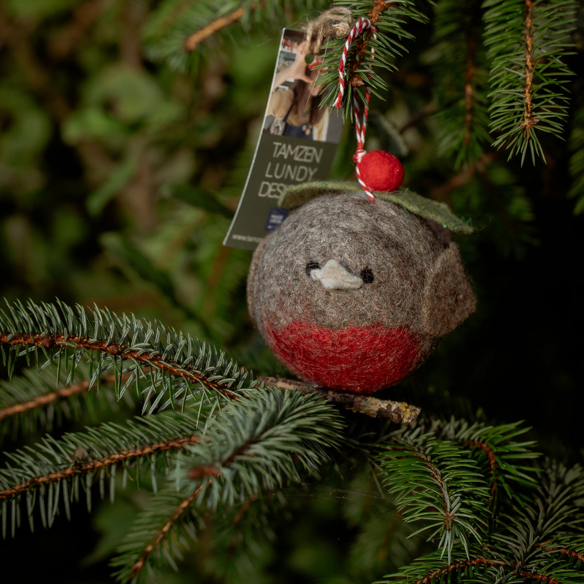 A felted bird Christmas ornament with a gray body, red belly, and green hat, hanging on a pine branch with its product tag still attached.