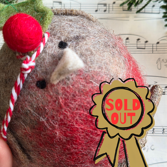 A close-up of a handmade felted wool bird decoration, likely a robin, with a red felted wool beak, black felted wool eyes, a white felted wool beak tip, a green felted wool hat, and a red and white striped cord. A gold 'SOLD OUT' ribbon graphic is overlaid on the bottom right.