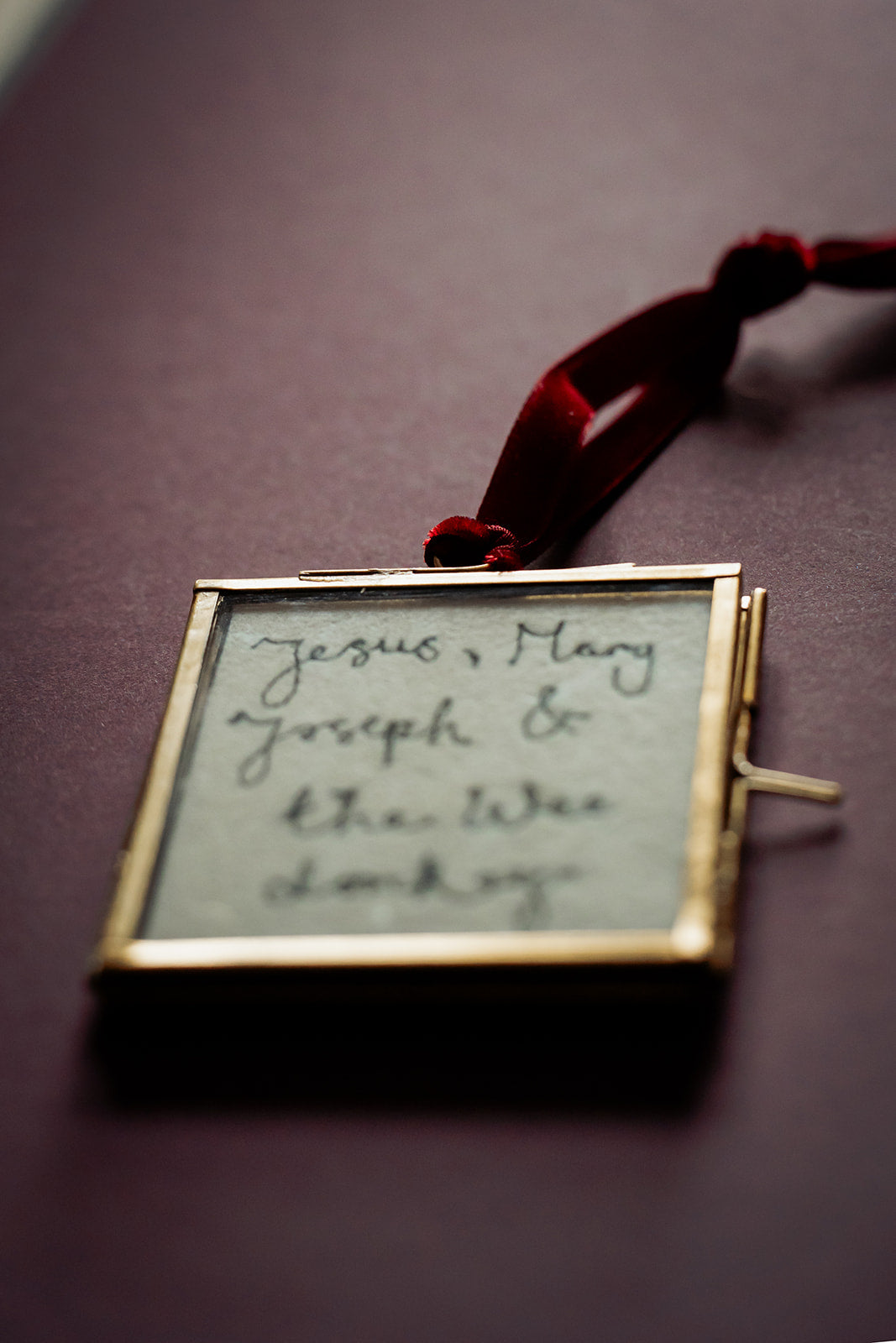 A square gold-framed keepsake box pendant with a red ribbon, containing handwritten text on a light background, shown on a dark purple surface.