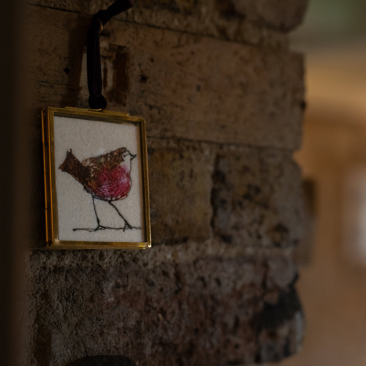 A small framed embroidered robin ornament hanging on a rustic brick wall. The ornament features a detailed black and white embroidery of a robin with a pink chest, enclosed in a gold frame and suspended by a black ribbon.
