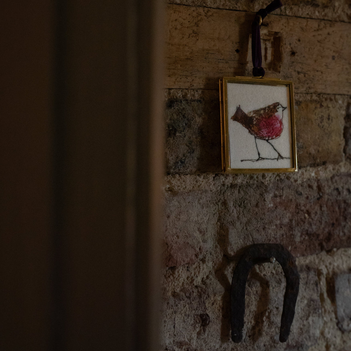 A small framed artwork featuring a robin hanging on a rustic brick wall, alongside a dark metal horseshoe, with a dark vertical foreground element on the left.