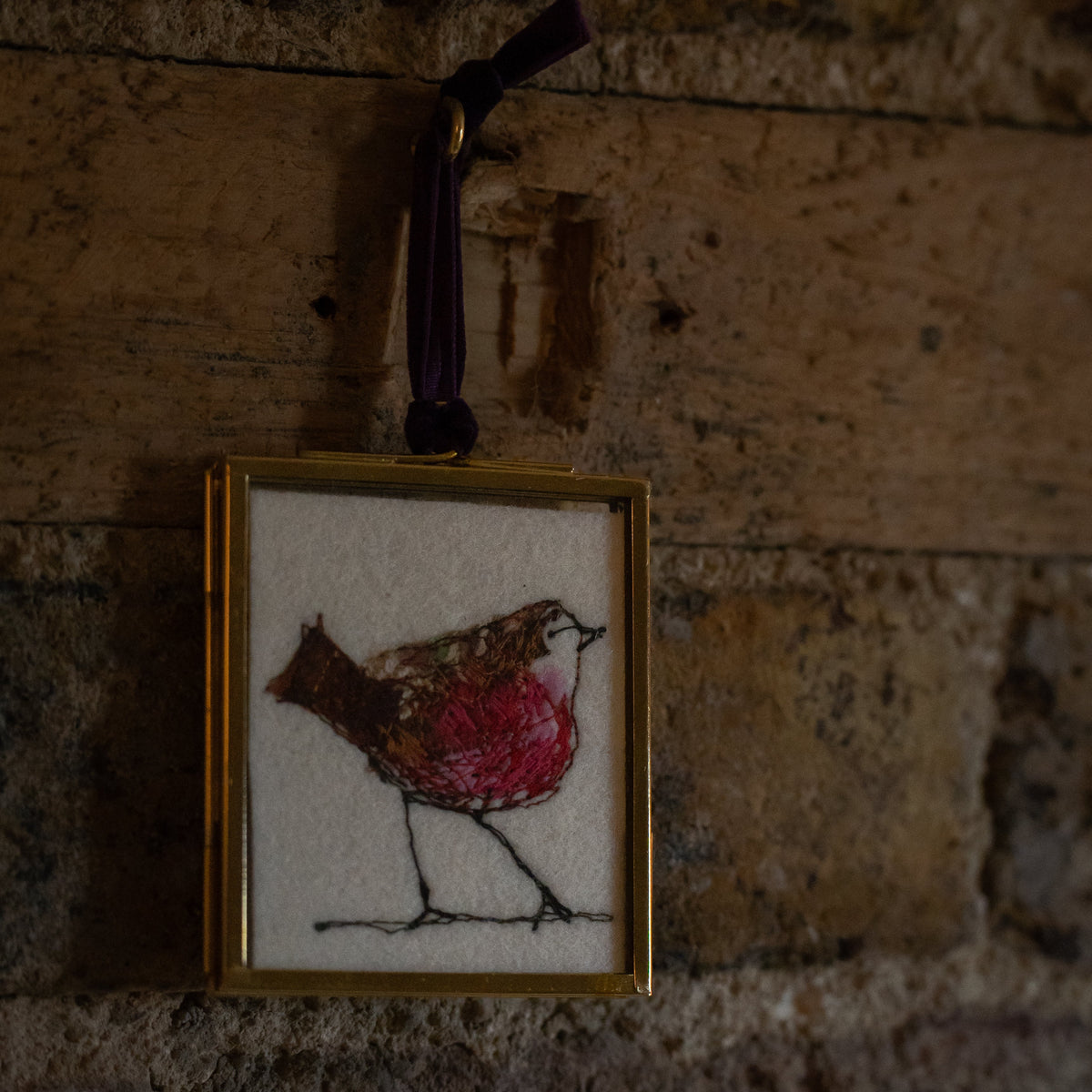 A small framed hanging ornament featuring a bird illustration with a red breast on a white background, suspended by a dark ribbon against a rustic wooden wall.