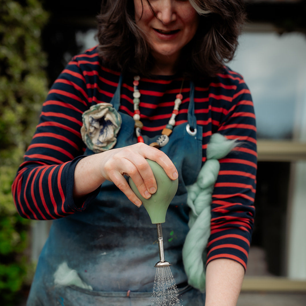 A woman wearing a red and navy striped shirt and a blue paint-splattered apron uses a green hand-pump bottle to spray water onto a woven mat on a wooden table.