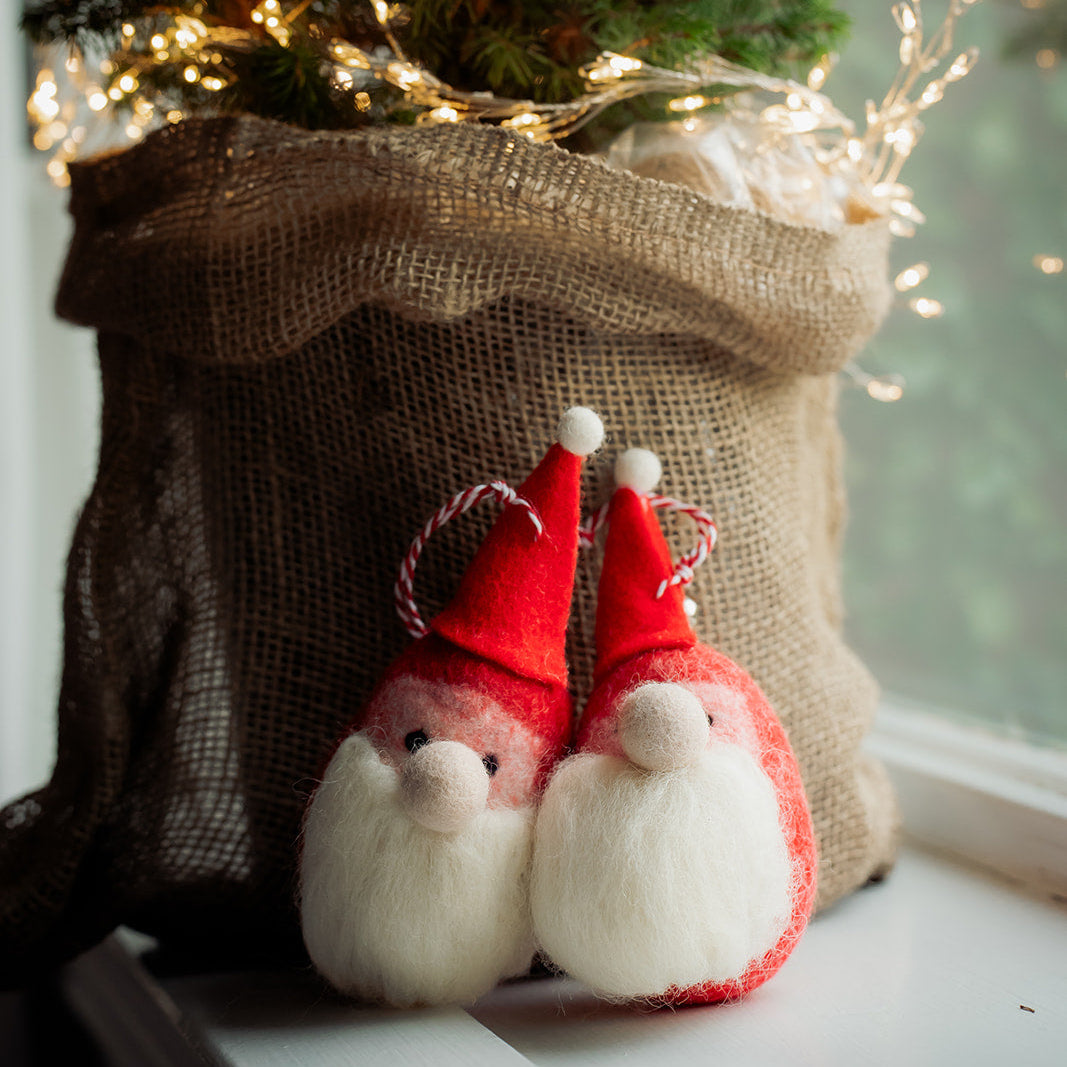 Two small red and white gnome ornaments with white beards sit in front of a burlap sack filled with evergreen branches and warm white fairy lights, on a windowsill.