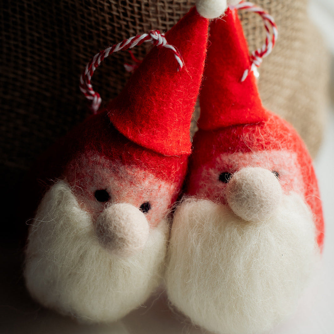 Two small felt Christmas gnome ornaments with red hats, white beards, and red-and-white hanging twine, posed in front of a burlap sack on a white surface.
