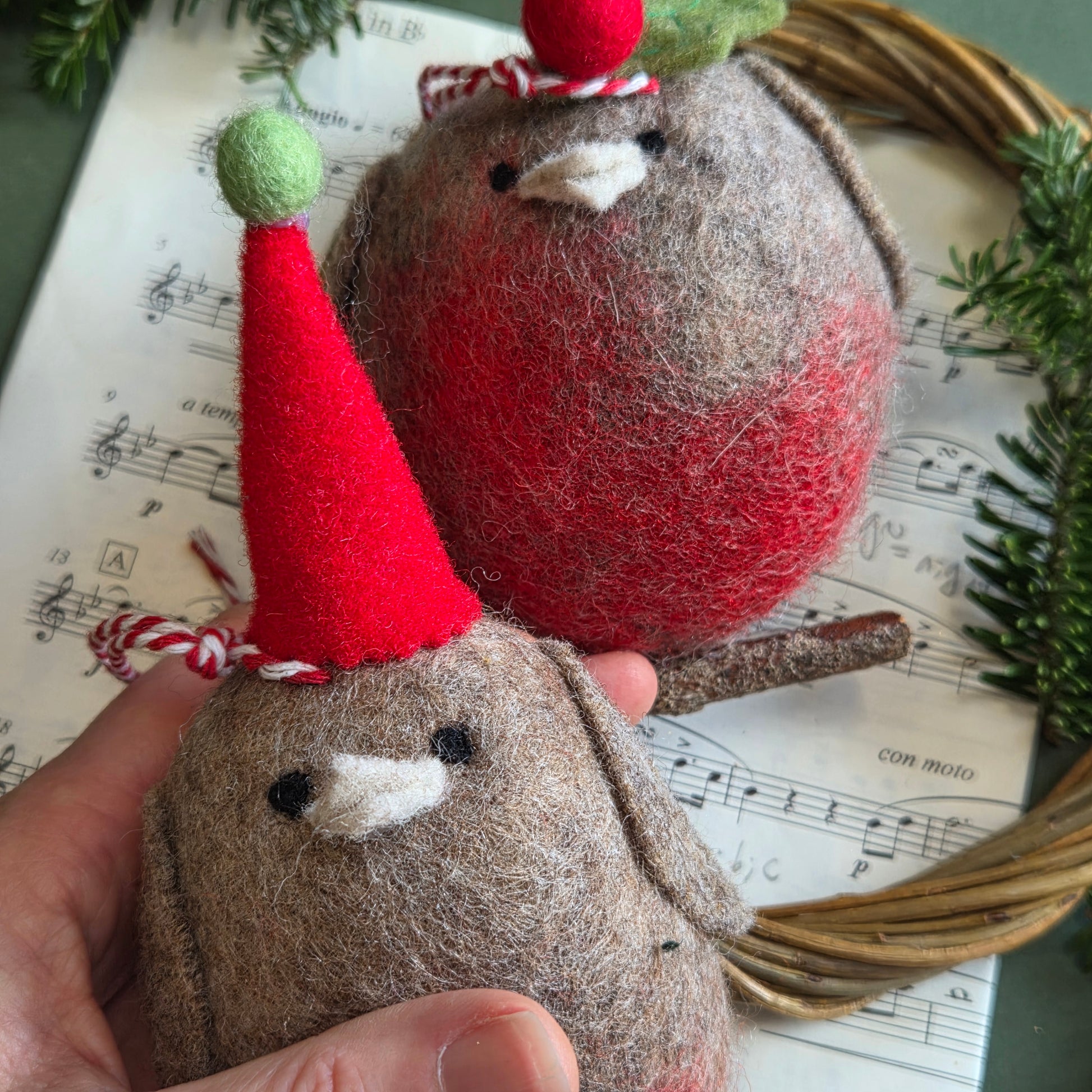 Two handmade felt Christmas ornaments shaped like birds, held in a hand. One ornament is red and gray with a green and red hat, and the other is gray with a red hat.