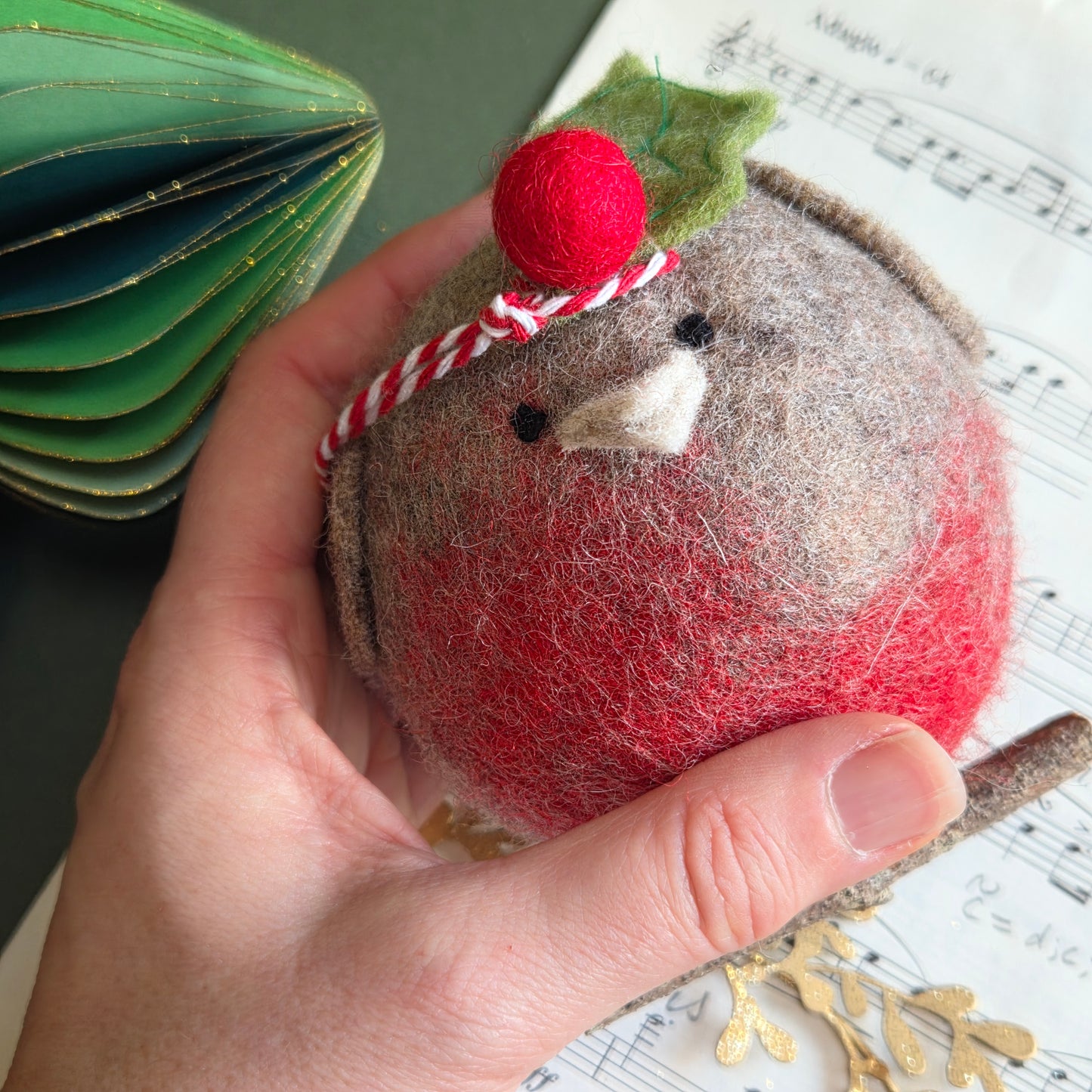 A hand holding a handmade felt Christmas ornament shaped like a bird, with red and brown felt, a small green leaf, a red pom-pom, and a red-and-white striped bow, against a background of sheet music.