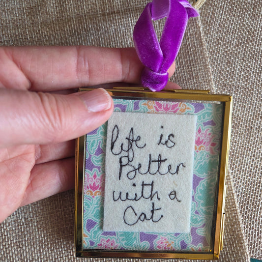 Small embroidered sign with 'Life is better with a cat' text held by a hand on a textured surface.