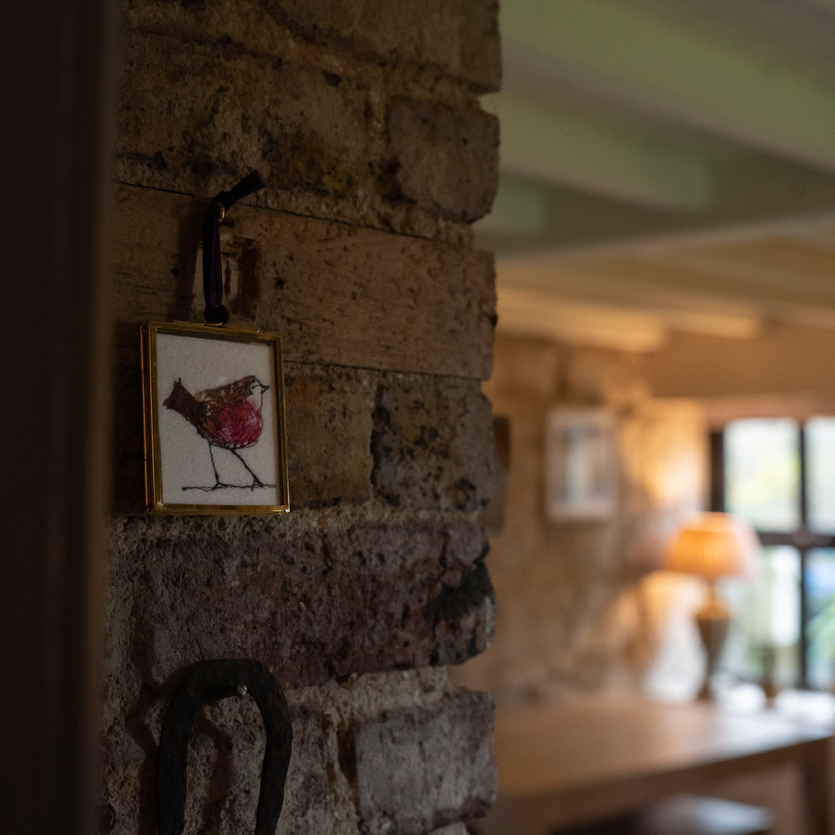 A small framed artwork featuring a bird hanging on a rustic stone wall in a warmly lit interior room.